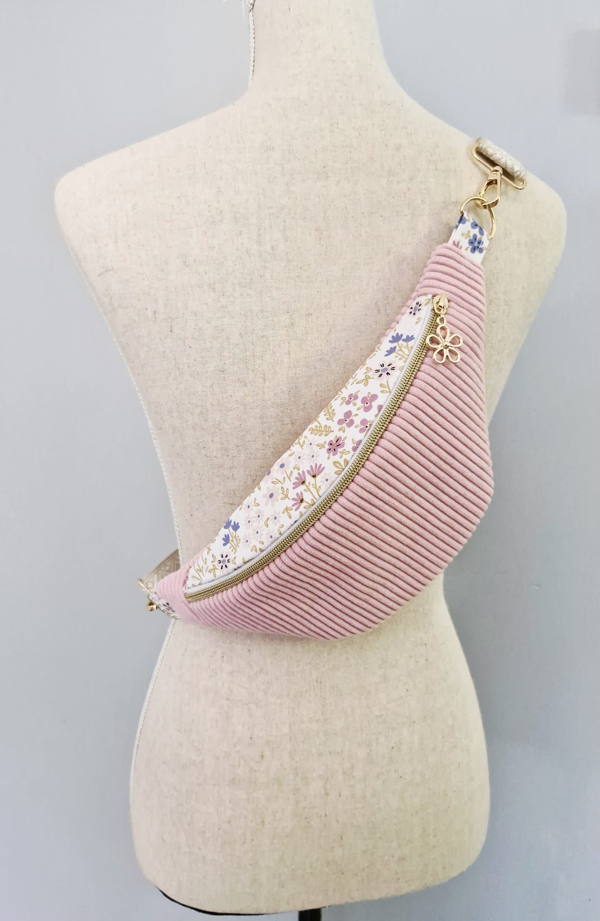Sac banane fait main en velours côtelé rose bonbon avec bande supérieure à motif fleuri, sangle beige réglable et pendentif doré, présenté sur mannequin et à plat.