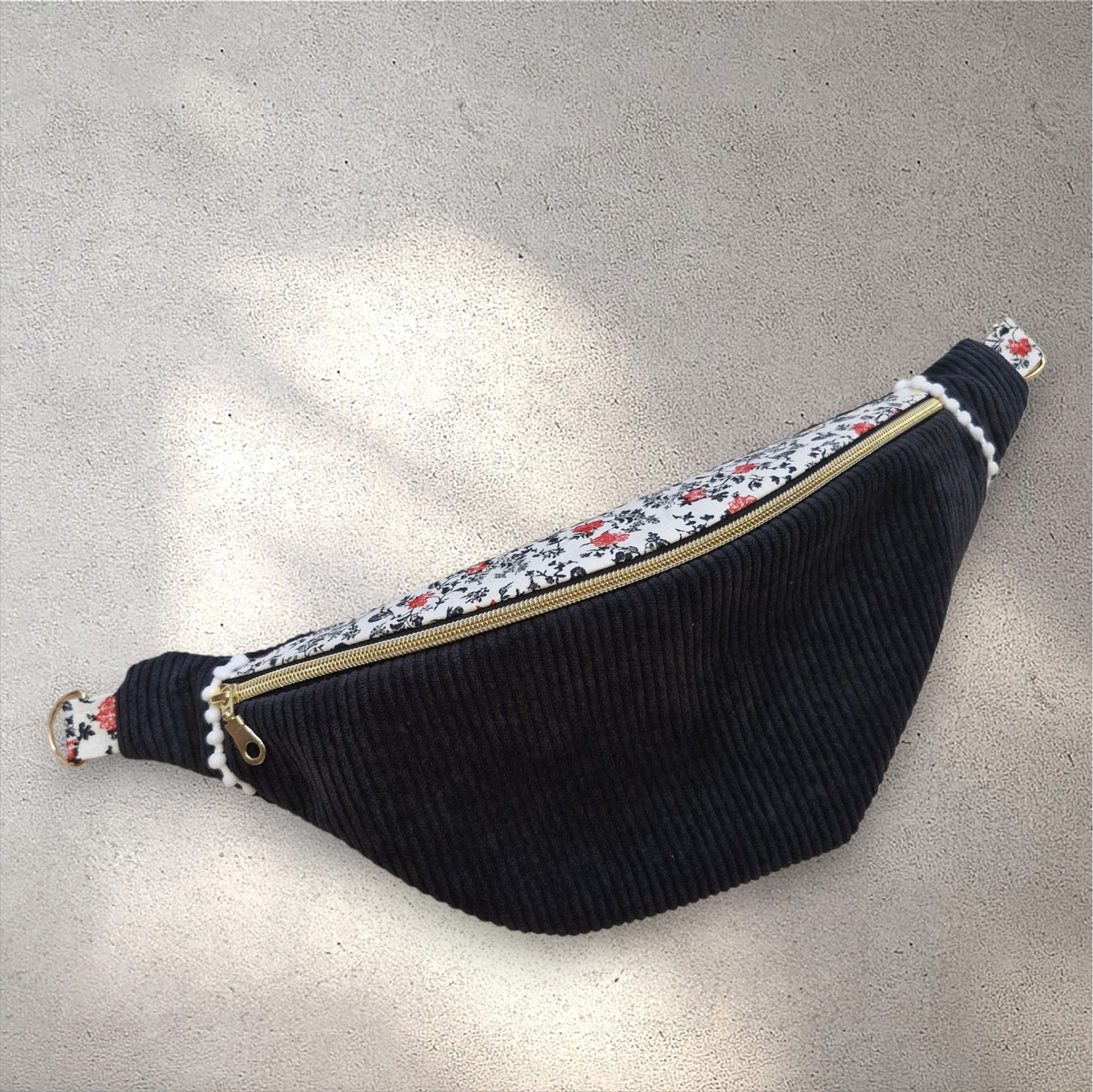 Sac banane fait main en velours côtelé noir avec rabat en tissu fleuri blanc, noir et rouge, zip doré, posé sur un fond en fausse fourrure beige. Sangle large amovible à motifs géométriques noirs et blancs, placée au-dessus du sac.