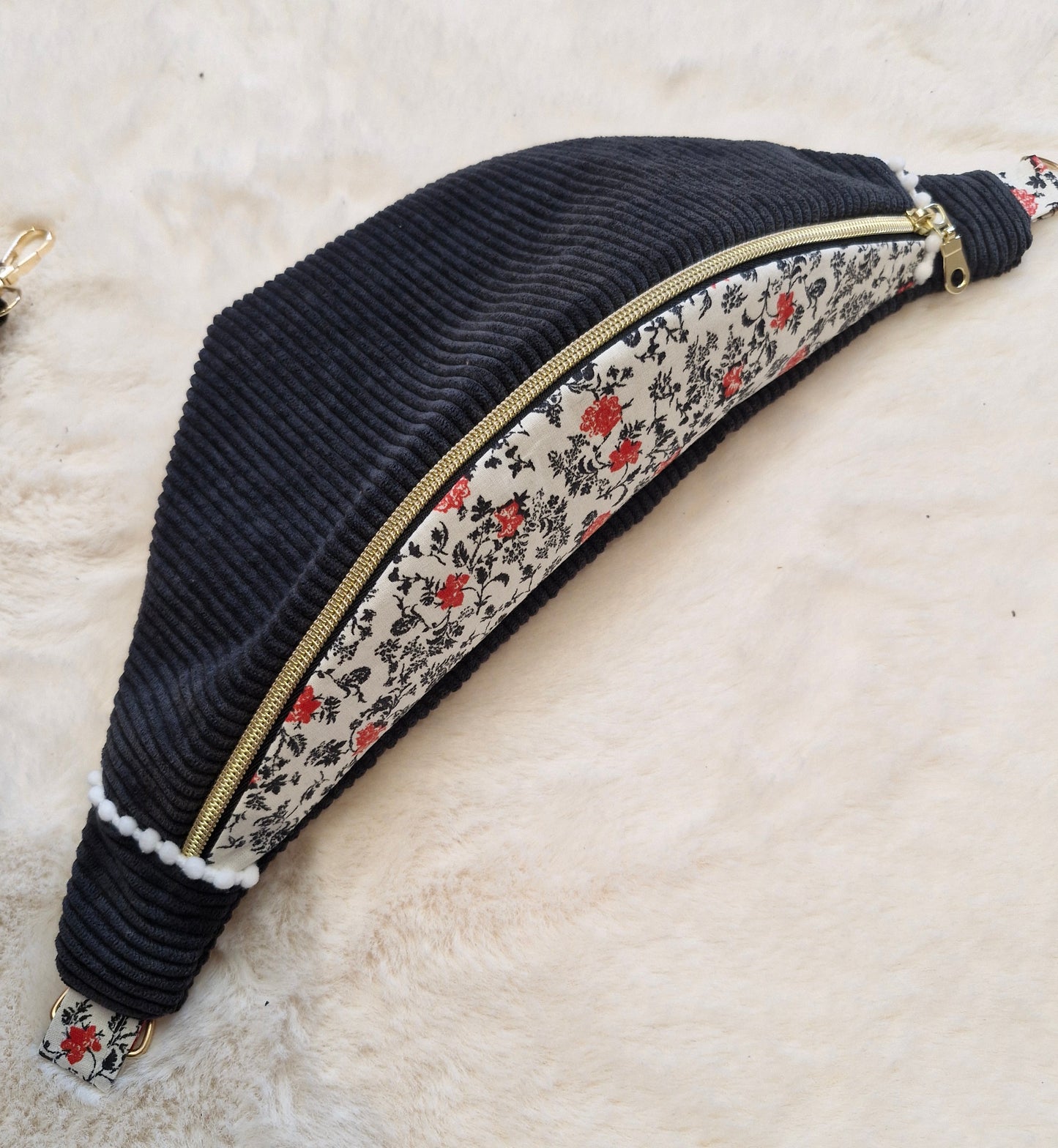 Sac banane fait main en velours côtelé noir avec rabat en tissu fleuri blanc, noir et rouge, zip doré, posé sur un fond en fausse fourrure beige. Sangle large amovible à motifs géométriques noirs et blancs, placée au-dessus du sac.