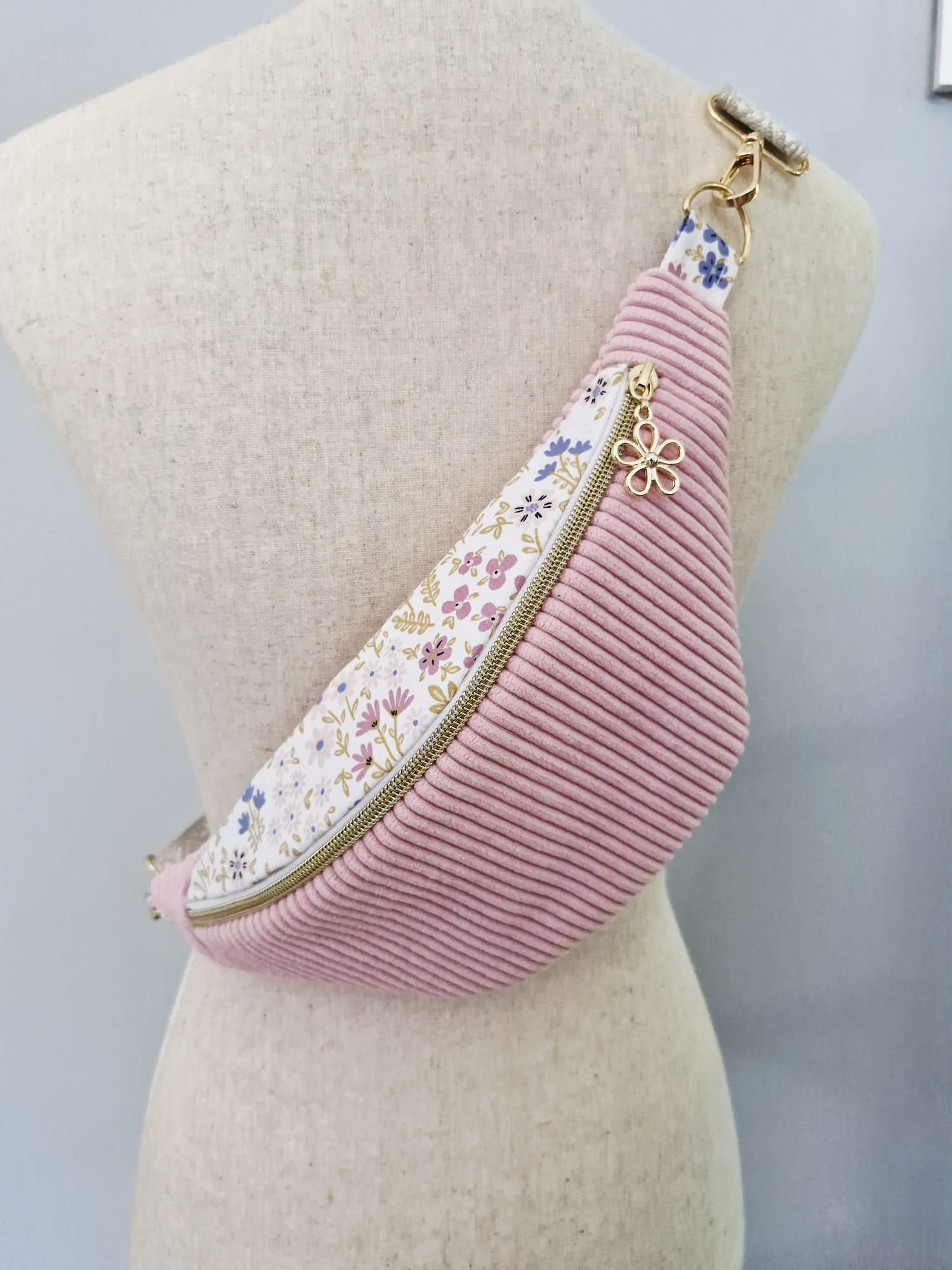 Sac banane fait main en velours côtelé rose bonbon avec bande supérieure à motif fleuri, sangle beige réglable et pendentif doré, présenté sur mannequin et à plat.