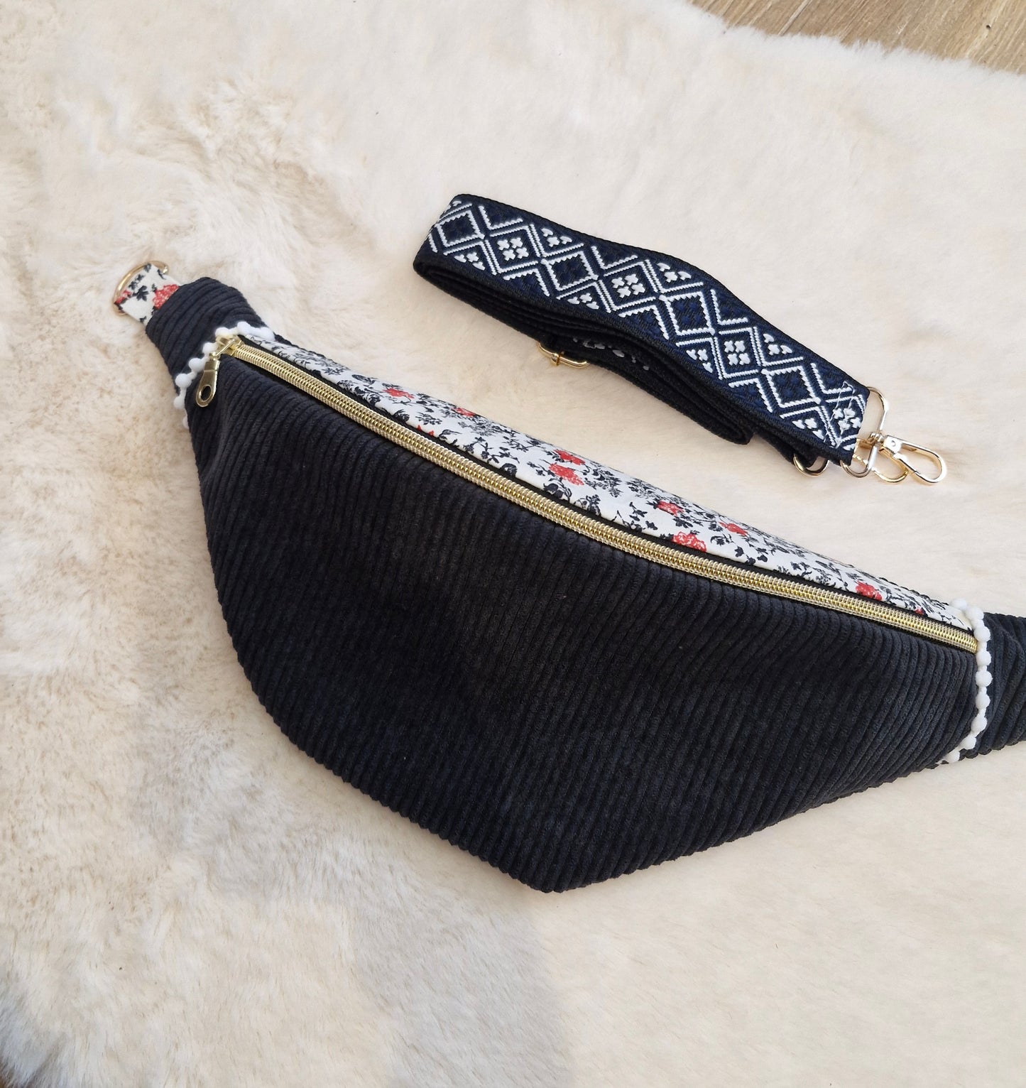 Sac banane fait main en velours côtelé noir avec rabat en tissu fleuri blanc, noir et rouge, zip doré, posé sur un fond en fausse fourrure beige. Sangle large amovible à motifs géométriques noirs et blancs, placée au-dessus du sac.